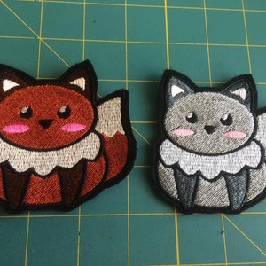 May include: Two embroidered iron-on patches of foxes. One fox is red with a white belly and tail tip. The other fox is gray with a white belly and tail tip.