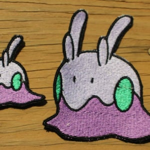 May include: Two purple and green embroidered patches of a cartoon creature with bunny ears. The larger patch is about 4 inches wide and the smaller patch is about 2 inches wide.