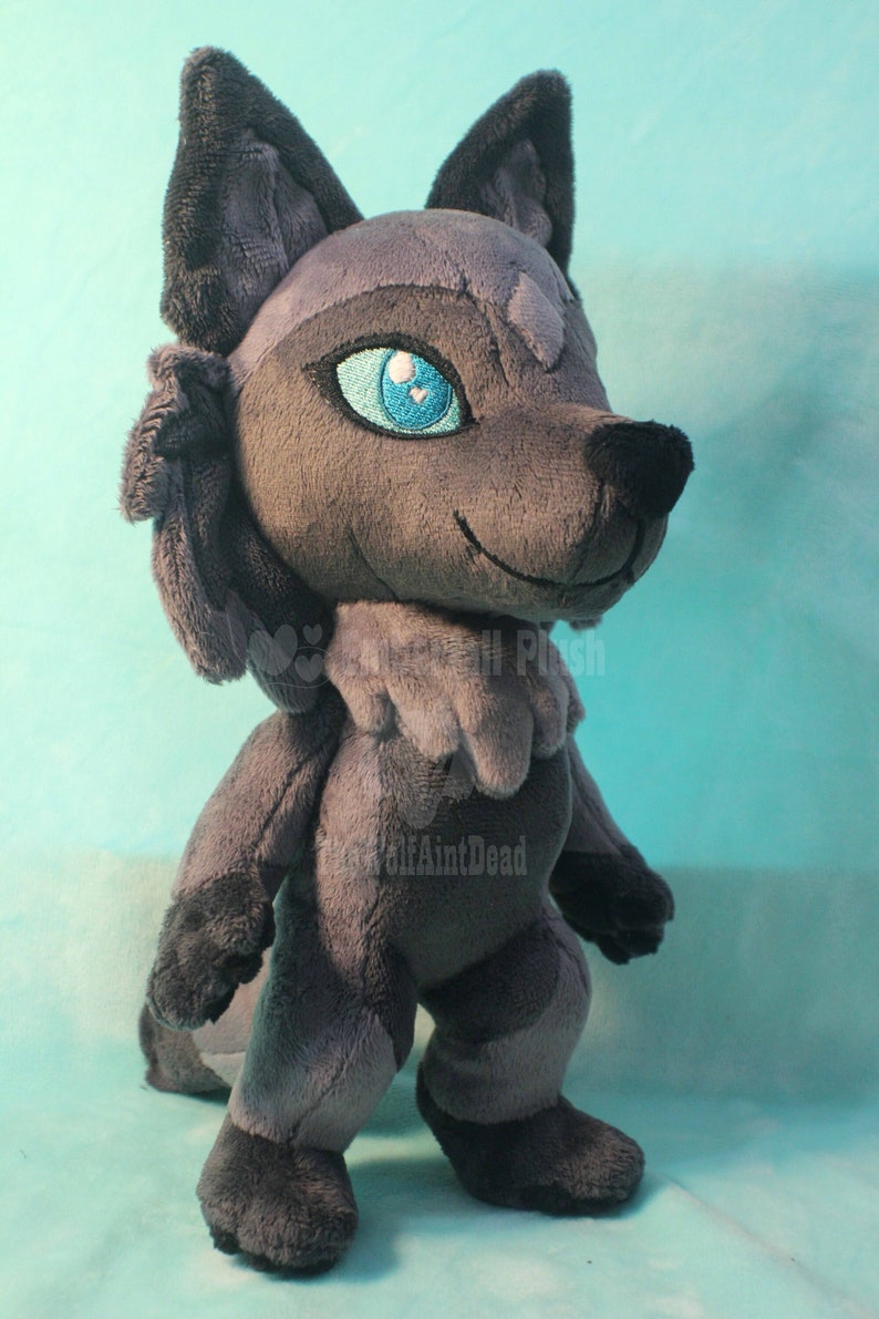 May include: A gray plush wolf with blue eyes. The wolf is standing on a light blue surface. The wolf has a fluffy tail and a small, white heart on its chest.