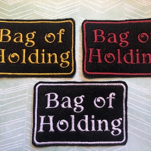 Peut inclure: Trois patchs en tissu noir avec des broderies blanches, dorées et rouges. Chaque patch porte l'inscription "Bag of Holding".