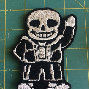 May include: A black and white embroidered patch of a skeleton character with a white skull and a grin. The character is wearing a black jacket and blue jeans. The character is waving with its right hand.