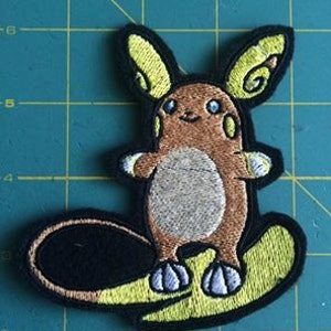 May include: A brown and yellow embroidered patch of a cartoon character surfing on a yellow surfboard. The character has large ears and a small mouth.