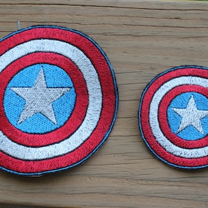 May include: Two embroidered patches with a shield design. Each patch has a white star on a blue circle, encircled by white and red rings. The patches are circular, one larger than the other. They are displayed on a wooden surface.