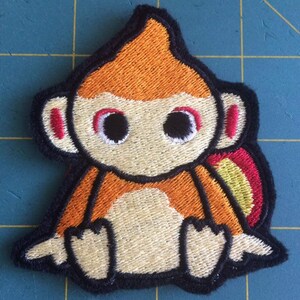 May include: Embroidered patch of a cartoon monkey with orange fur, a yellow and red tail, and black eyes. The monkey is sitting with its legs crossed and has a white belly.