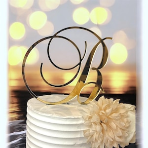 May include: A gold-colored letter "B" cake topper in a cursive font, placed on a white frosted cake. A cream-colored flower is on the side of the cake. The background is blurred with warm tones.