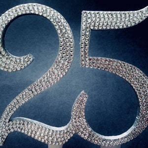 Peut inclure: Le chiffre 25 de couleur argenté, composé de strass. Le chiffre est sur un fond bleu foncé.