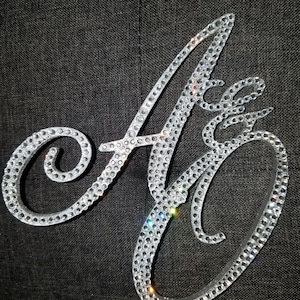 May include: A silver cake topper featuring the letters "A & G" encrusted with sparkling rhinestones. The letters are in a cursive font and are attached to a silver base. The background is a dark gray fabric.
