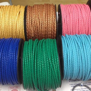 May include: A collection of spools of braided cord in various colors, including black, gold, bronze, pink, red, gray, blue, green, light blue, and orange. The cords are tightly wound on black spools.