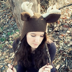 May include: A brown crocheted deer hat with white antlers. The hat has ear flaps and is worn by a person with long brown hair.