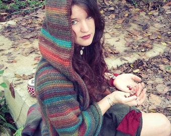 May include: A woman wearing a multi-colored striped crocheted hooded cardigan with a dark green skirt and brown boots. She is holding a small red pouch in her hand.