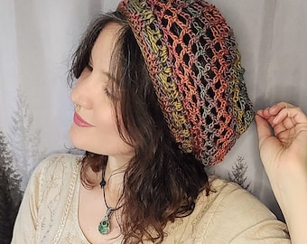 May include: A woman wearing a crocheted beanie hat in shades of brown, orange, and gray. The hat has a loose, open weave and is worn on top of her head. She is wearing a cream-colored, lace-trimmed top with a tied front and blue jeans.