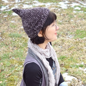 May include: A woman wearing a purple and gray knitted hat with a pointed top, a gray and white lace scarf, a gray vest, and gray and black pants. She is sitting on a grassy surface with snow in the background.