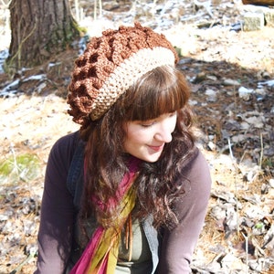 May include: A woman wearing a brown and beige crocheted beanie with a long, multi-coloured scarf and a green dress. She is sitting on a mossy rock in a wooded area.