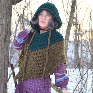 May include: A woman wearing a green and brown crocheted hooded shawl with a purple and white patterned dress. She is holding a green wreath in her hand. The background is a snowy woodland.