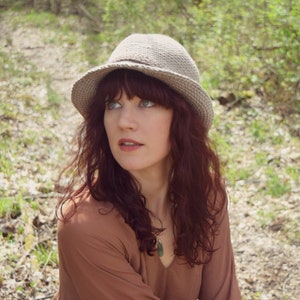 May include: A woman wearing a brown dress and a gray fedora hat with a wide brim. She is sitting in a forest setting with green foliage and brown dirt.