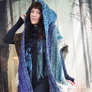 May include: A hooded crocheted cardigan with a blue, purple, and white color gradient. The cardigan has a large hood and a loose fit. The person wearing the cardigan is also wearing black jeans and boots.