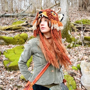 Könnte beinhalten: Eine Frau trägt eine braune und orangefarbene, gestrickte Mütze in Form eines Fuchses mit langen Quasten, eine grüne Jacke, eine braune Ledertasche und einen Patchworkrock.