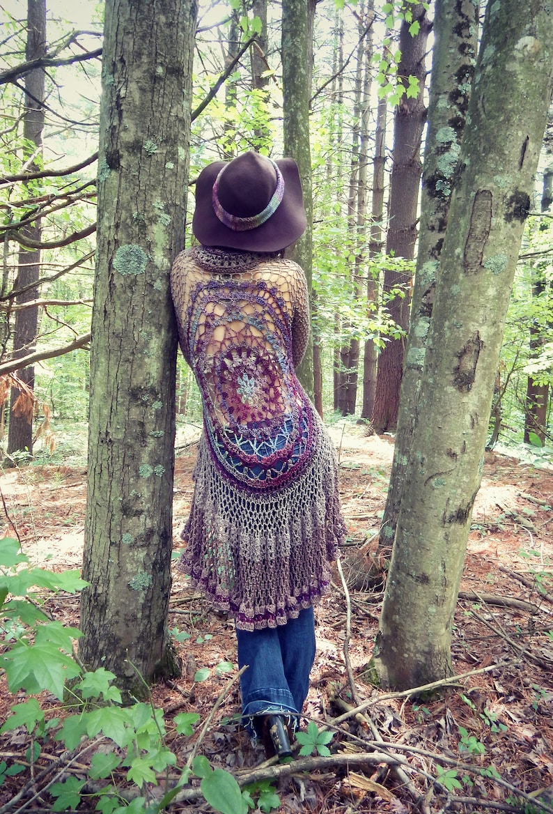 Pu&ograve; includere: Una donna indossa un cardigan di uncinetto marrone con un design a mandala colorato, un cappello di feltro marrone e jeans blu e si trova in un ambiente boschivo.