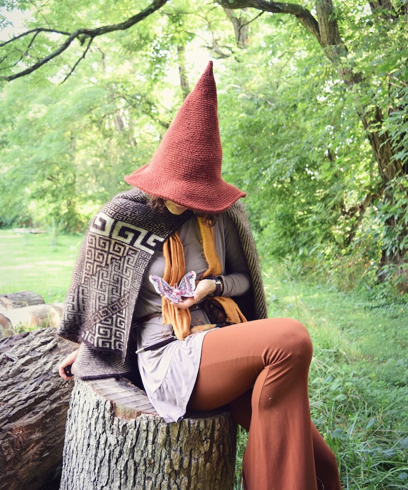 May include: A person wearing a brown pointed hat with a geometric patterned shawl and brown pants is sitting on a tree stump holding a butterfly.