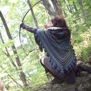 Puede incluir: Una mujer en un entorno de bosque, con un chal de ganchillo con capucha con un patr&oacute;n de rayas multicolores, sostiene un arco y una flecha, apuntando a un objetivo invisible.