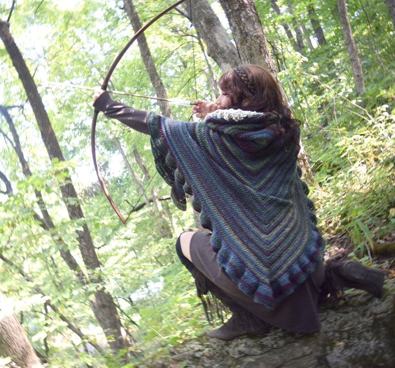Puede incluir: Una mujer en un entorno de bosque, con un chal de ganchillo con capucha con un patr&oacute;n de rayas multicolores, sostiene un arco y una flecha, apuntando a un objetivo invisible.