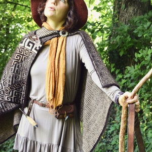 May include: A woman wearing a brown knitted cape with a geometric pattern, a brown hat with a pointed top, a brown leather belt, and a long brown scarf. She is holding a wooden staff with a leather strap.