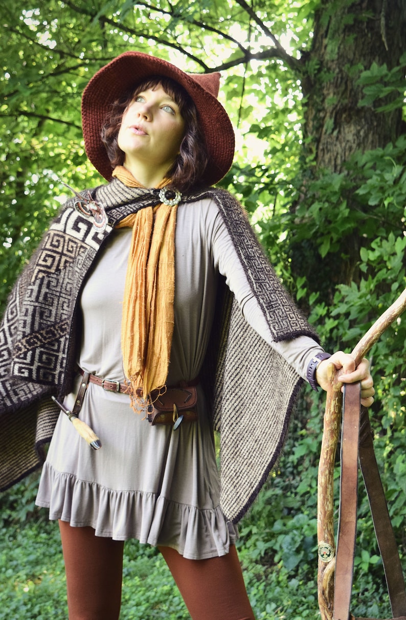 May include: A woman wearing a brown knitted cape with a geometric pattern, a brown hat with a pointed top, a brown leather belt, and a long brown scarf. She is holding a wooden staff with a leather strap.
