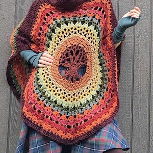 Puede incluir: Una mujer lleva un poncho de ganchillo con un diseño de árbol de la vida en tonos de marrón, naranja, amarillo, verde y gris. El poncho tiene un cuello alto y se lleva sobre una falda de cuadros y botas marrones.