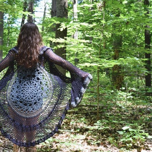 Pu&ograve; includere: Un cardigan in crochet nero e grigio con maniche a campana. Il cardigan &egrave; indossato sopra un top in crochet grigio e bianco. La persona &egrave; in piedi in un bosco.