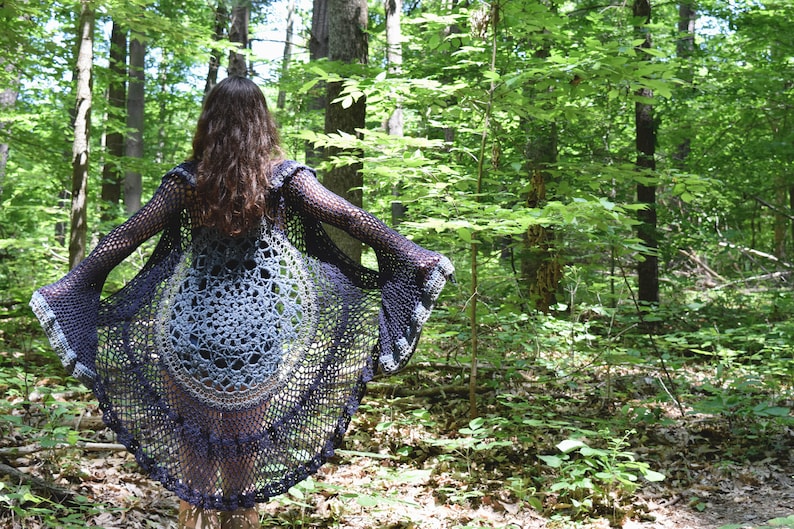 Pu&ograve; includere: Un cardigan in crochet nero e grigio con maniche a campana. Il cardigan &egrave; indossato sopra un top in crochet grigio e bianco. La persona &egrave; in piedi in un bosco.