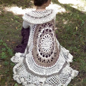 Pu&ograve; includere: Un cardigan bianco all'uncinetto con un design circolare marrone e bianco sul retro. Il cardigan ha un ampio colletto all'uncinetto e un orlo arricciato.