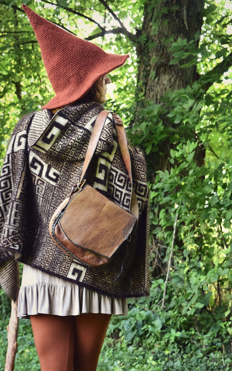 May include: A person wearing a brown and white patterned poncho with a brown leather satchel and a red pointed hat. The person is standing in a forest setting.