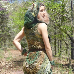 Pu&ograve; includere: Una donna indossa un abito di uncinetto verde e marrone con un top con cappuccio abbinato. L'abito ha una gonna fluida e un corpetto aderente. La donna &egrave; in piedi in un ambiente boschivo.