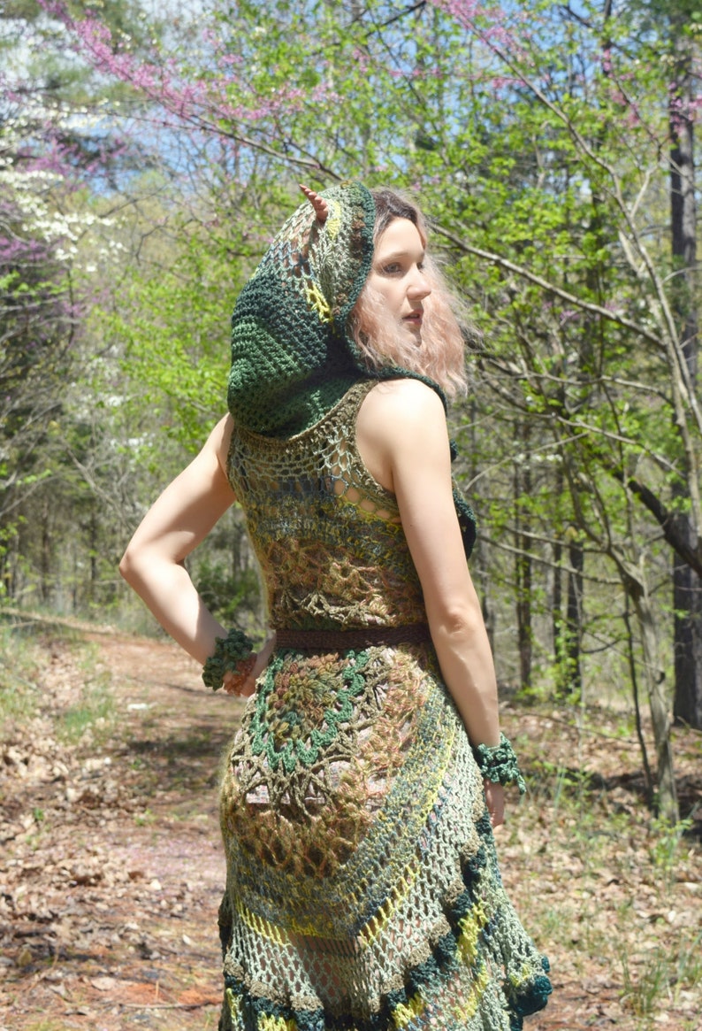 Pu&ograve; includere: Una donna indossa un abito di uncinetto verde e marrone con un top con cappuccio abbinato. L'abito ha una gonna fluida e un corpetto aderente. La donna &egrave; in piedi in un ambiente boschivo.