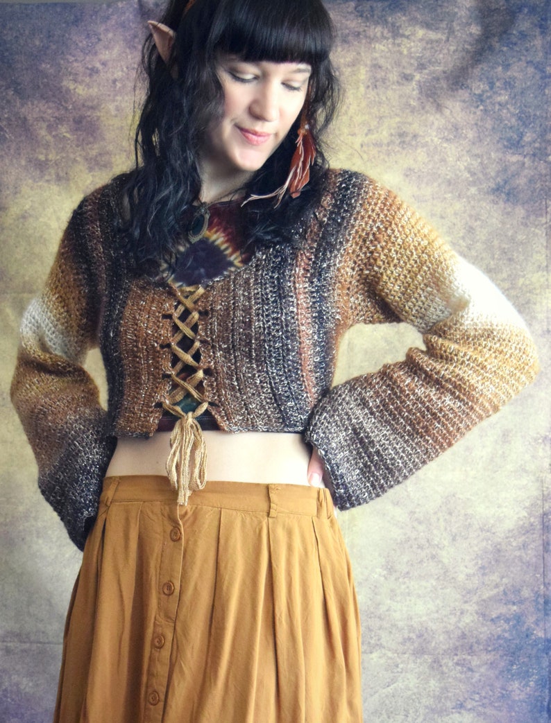May include: A brown and white crocheted sweater with a lace-up front and long sleeves. The sweater is cropped and worn with a mustard yellow skirt.