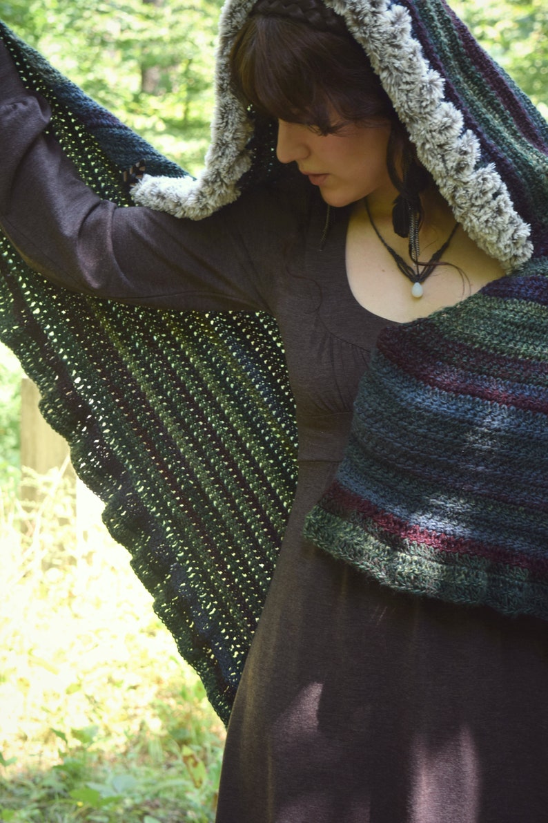 Puede incluir: Una mujer con un vestido marr&oacute;n y una capa con capucha. La capa est&aacute; hecha a ganchillo con un patr&oacute;n de rayas verdes, azules y moradas. La capucha tiene un borde de piel blanca.