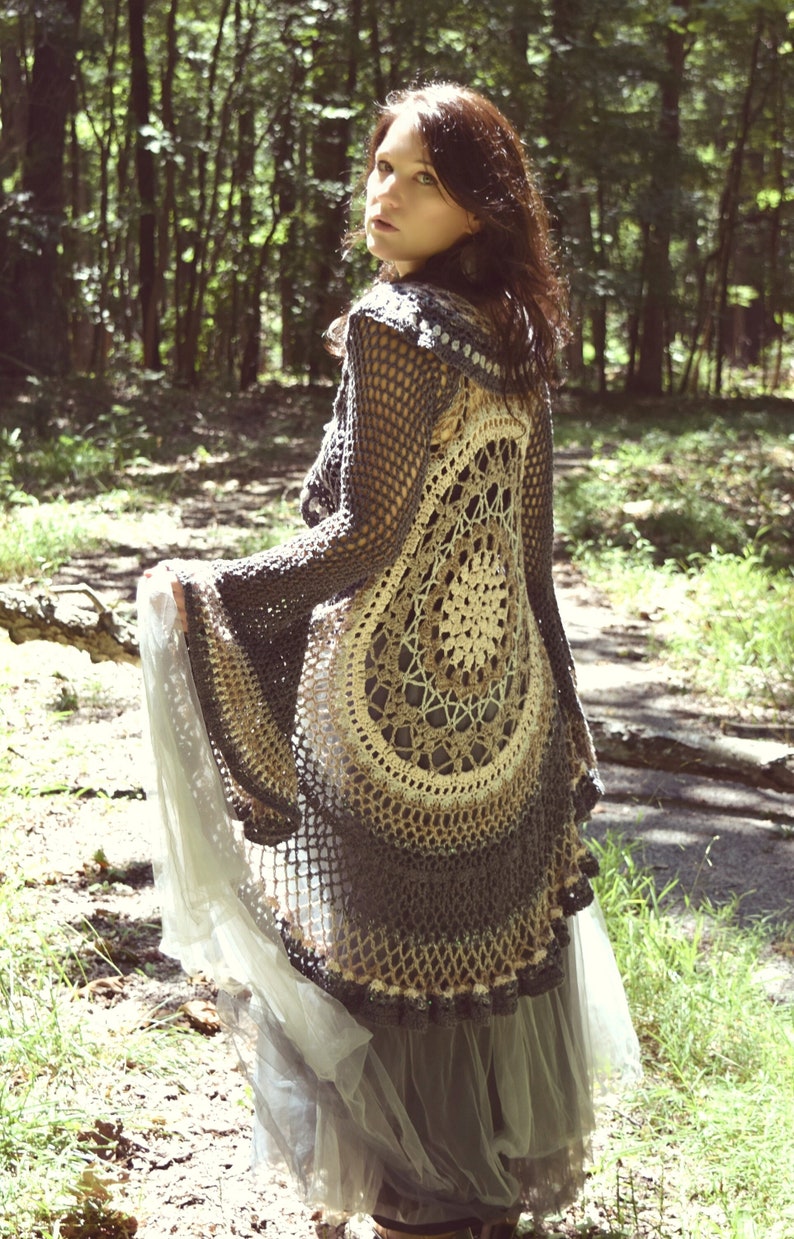 Pu&ograve; includere: Una donna indossa un cardigan all'uncinetto con un grande disegno circolare sul retro. Il cardigan &egrave; grigio e beige e ha uno stile fluido e boh&eacute;mien. La donna &egrave; in piedi in un ambiente boschivo.