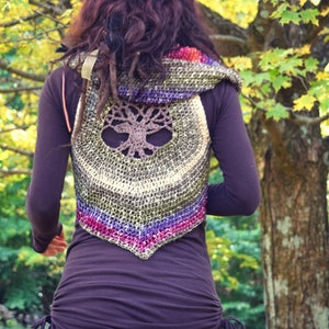 May include: A woman wearing a brown crocheted vest with a tree of life design. The vest has a hood and is made with a variety of colors, including green, purple, and pink. The woman is wearing a brown shirt and has long brown hair.