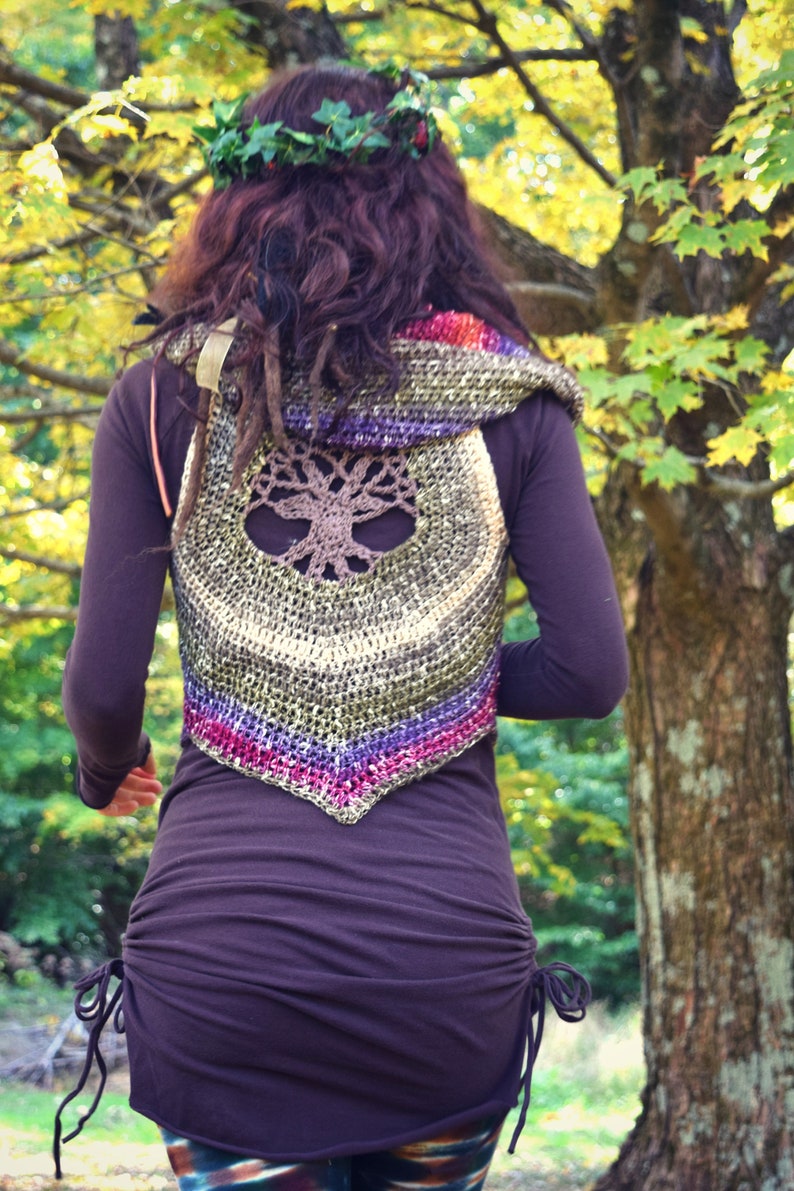 May include: A woman wearing a brown crocheted vest with a tree of life design. The vest has a hood and is made with a variety of colors, including green, purple, and pink. The woman is wearing a brown shirt and has long brown hair.