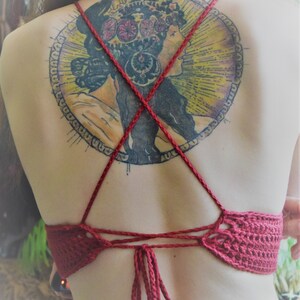 May include: A red crocheted halter top with a back tie closure. The top is worn by a person with a large circular tattoo on their back. The tattoo features a woman with a floral crown and a colorful design.