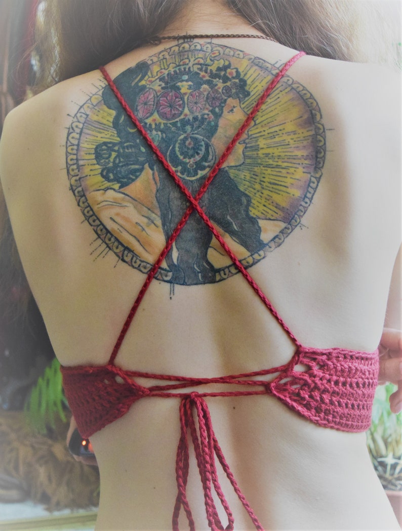 May include: A red crocheted halter top with a back tie closure. The top is worn by a person with a large circular tattoo on their back. The tattoo features a woman with a floral crown and a colorful design.