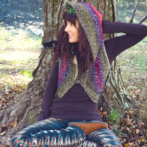 May include: A woman wearing a colorful crocheted hooded vest and tie-dye leggings sits in front of a tree. The vest has a rainbow pattern and the leggings have a blue, green, and brown pattern.