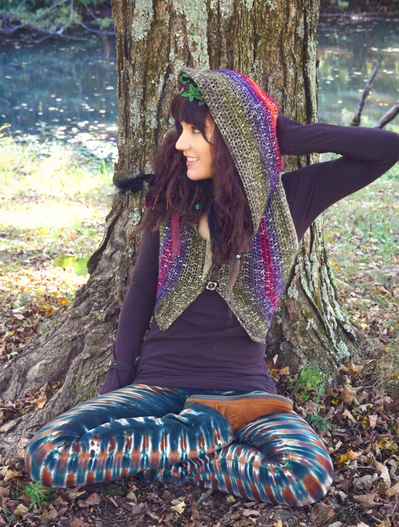 May include: A woman wearing a colorful crocheted hooded vest and tie-dye leggings sits in front of a tree. The vest has a rainbow pattern and the leggings have a blue, green, and brown pattern.