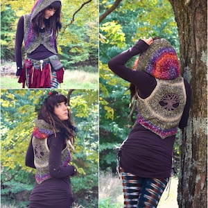 May include: A woman wearing a crocheted vest with a tree of life design. The vest has a rainbow colored hood and is worn over a brown shirt. The woman is standing in a forest setting.