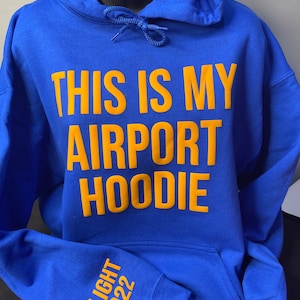 May include: A royal blue hoodie with the text "THIS IS MY AIRPORT HOODIE" in yellow lettering. The left sleeve has the text "FLIGHT 1922" in yellow lettering.