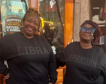 Op de afbeelding: Twee mensen dragen zwarte sweatshirts met hun sterrenbeelden, Weegschaal en Steenbok, in wit gedrukt. De persoon links draagt ​​een blauwe jeans en de persoon rechts draagt ​​een blauwe jeans.