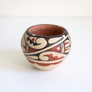May include: A small, round, terracotta pot with a geometric design in black, red, and white. The pot is decorated with a pattern of swirls, zigzags, and dots.