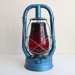 May include: A vintage blue lantern with a red glass globe. The lantern has a metal frame and a handle for carrying. The lantern is approximately 12 inches tall.