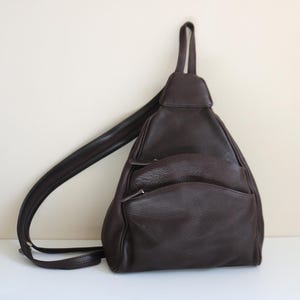May include: A dark brown leather sling backpack with a single shoulder strap. The bag has a triangular shape with two zippered front pockets and a top handle. The leather has a textured appearance, and the bag is sitting on a white surface.