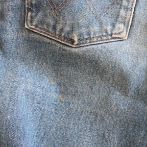 May include: Close-up of blue denim fabric with a back pocket. The fabric has a worn, faded look with visible stitching and a slight sheen.